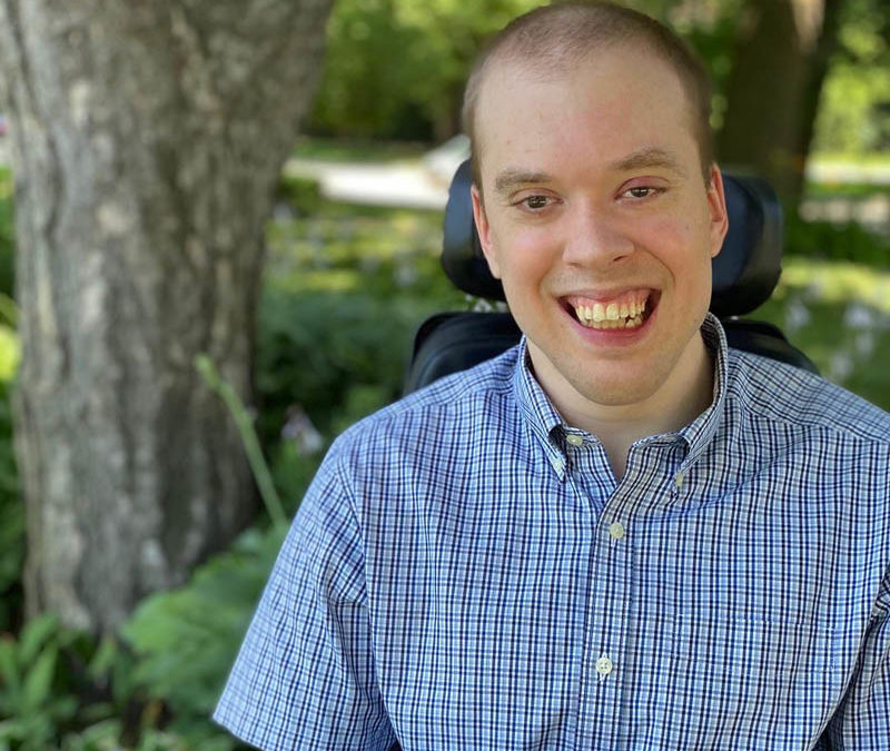 A photo of David Gayes, a smiling white man in a wheelchair and blue shirt. Trees are in the background.