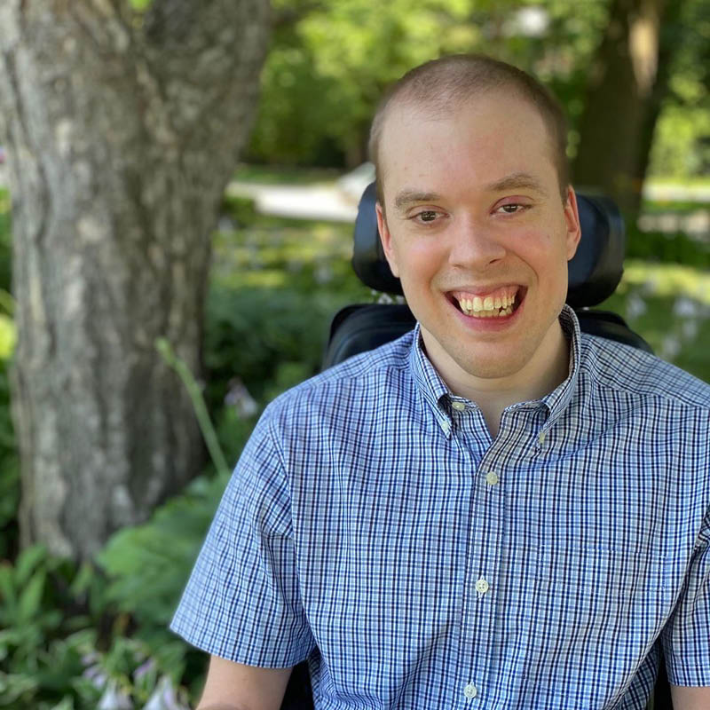 A photo of David Gayes, a smiling white man in a wheelchair and blue shirt. Trees are in the background.
